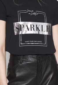 Camiseta negra con estampado metálico plateado que dice "SPARKLE" y "MAKE YOUR OWN MAGIC"; material de algodón suave, corte relajado, mangas cortas.