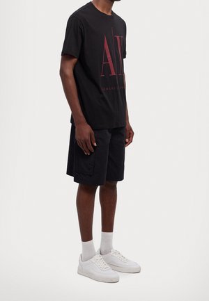T-shirt en coton noir avec un grand logo rouge "AE", associé à un short sombre et des baskets blanches. Design épuré, manches courtes, coupe décontractée.