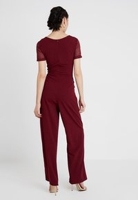 Burgundský overal s krátkými krajkovými rukávy, přiléhavým živůtkem a širokými nohavicemi. Zapínání na zip na zadní straně. Hladká textura látky po celé ploše.