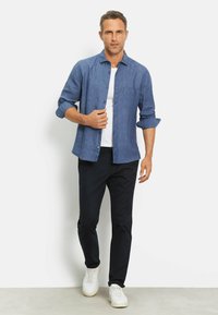 Blauw linnen shirt met een knoopsluiting aan de voorkant en een zak, gedragen over een wit T-shirt, gecombineerd met donkere broeken en witte sneakers.