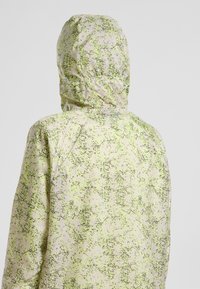 Veste à capuche en matériau léger, présentant un motif tacheté vert et noir sur un fond clair, avec une coupe décontractée.
