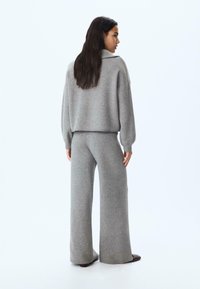 Svetlo siva pletena loungewear obleka, ki vključuje ohlapen pulover z visokim ovratnikom in široke hlače, oboje z mehko teksturo.