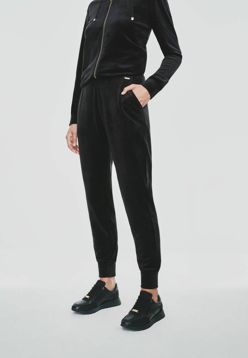 Osoba w czarnej welurowej kurtce z przeszyciem na zamek i dopasowanych spodniach dresowych, w czarnych trampkach, stoi z jedną ręką w kieszeni.