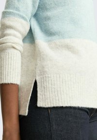 Pull rayé bleu clair et crème avec des poignets et un ourlet côtelés, fentes sur les côtés, en tissu doux et texturé. Porté sur un jean foncé.