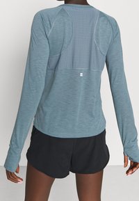 Langärmliges Sportoberteil in Hellblau, mit einem strukturierten Rückenpanel und Perforationen für Atmungsaktivität, kombiniert mit schwarzen Shorts.