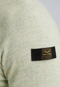 Hellgrüner Strickpullover mit strukturiertem Finish. Mit einem schwarzen Etikett mit goldener Schrift. Nahaufnahme des Schulterbereichs.