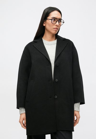 Čierny kabát zo zmesi vlny s výstrihom s výrezom, zapínaním na gombíky a bočnými vreckami. Nosený cez svetlosivý pletený sveter.