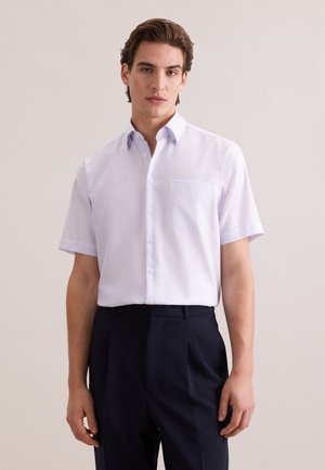 Uomo che indossa una camicia bianca a quadri a maniche corte con bottoni e pantaloni scuri plissettati, in piedi davanti a uno sfondo chiaro e uniforme.