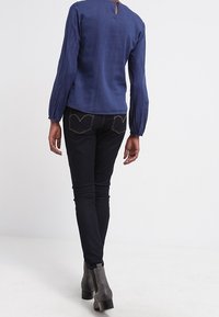 Person som går bort ifrån kameran, iklädd en blå långärmad blus, svarta åtsittande jeans och grå ankelstövlar mot en enkel vit bakgrund.