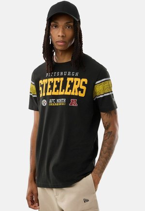 Man met een zwarte Pittsburgh Steelers T-shirt met gele strepen op de mouwen, beige broek en zwarte pet, staand tegen een effen achtergrond.