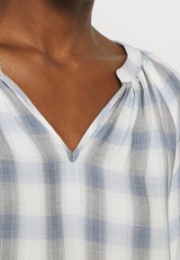 Blouse à carreaux bleus et blancs avec un décolleté en V et des détails de plissage délicats, fabriquée en tissu léger, montrant une texture douce.