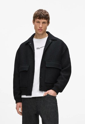 Junger Mann trägt eine schwarze, geknöpfte Jacke über einem weißen Karl Lagerfeld Jeans Hemd und dunklen Nadelstreifenhosen, steht mit einer Hand in der Tasche.