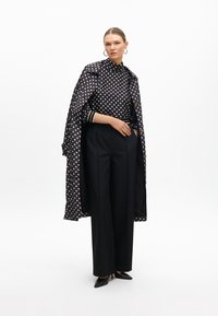 Schwarzes Polka-Dot-Hemd mit hohem Kragen, kombiniert mit hoch geschnittenen schwarzen Hosen und einem glänzenden, gemusterten Mantel. Die Ärmel zeigen Streifen. Schwarze High Heels.