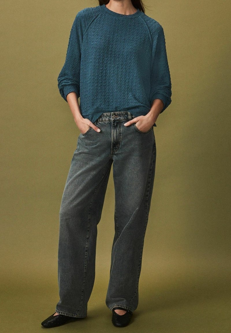 Personne portant une chemise bleue texturée à manches longues rentrée dans un jean noir délavé ample, les mains dans les poches et des chaussures noires, debout sur un sol olive.