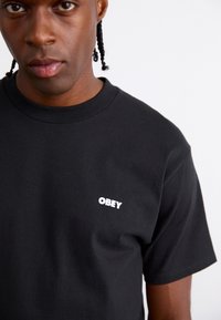 Zwarte T-shirt met korte mouwen van katoen, met een witte "OBEY"-logo op de linkerborst. Glad textuur, klassiek ronde hals ontwerp.