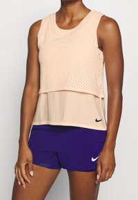 Canotta a rete color pesca con design a strati e logo nero, abbinata a pantaloncini sportivi viola. Tessuto leggero e traspirante.