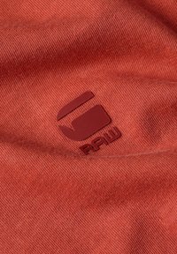 Tessuto rosso con una superficie liscia, caratterizzato da un logo in rilievo color bordeaux. Il design è semplice e mette in risalto il nome del marchio "RAW."