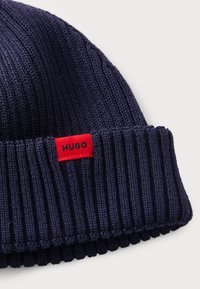 Marineblå strikket lue med ripet tekstur, med et r�ødt stoffmerke merket "HUGO." Designet er brettet ved kanten for en tettsittende passform.
