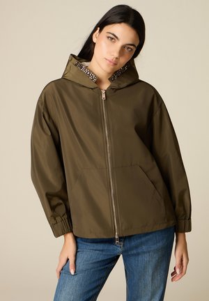 Frau trägt eine olivgrüne Jacke mit Reißverschluss, einer Kapuzenfütterung mit Perlen und blauen Jeans, steht vor einem einfarbigen beigen Hintergrund.