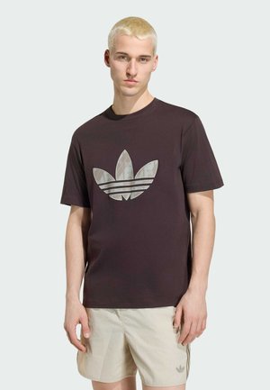 Mladý muž krátkými blond vlasy, který nosí černé tričko Adidas s velkým logem a béžové šortky s malým logem.