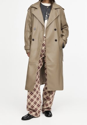 Personne portant un long manteau beige croisé sur un pull sombre et une chemise blanche, associé à un pantalon large beige et marron à carreaux et des chaussures noires.