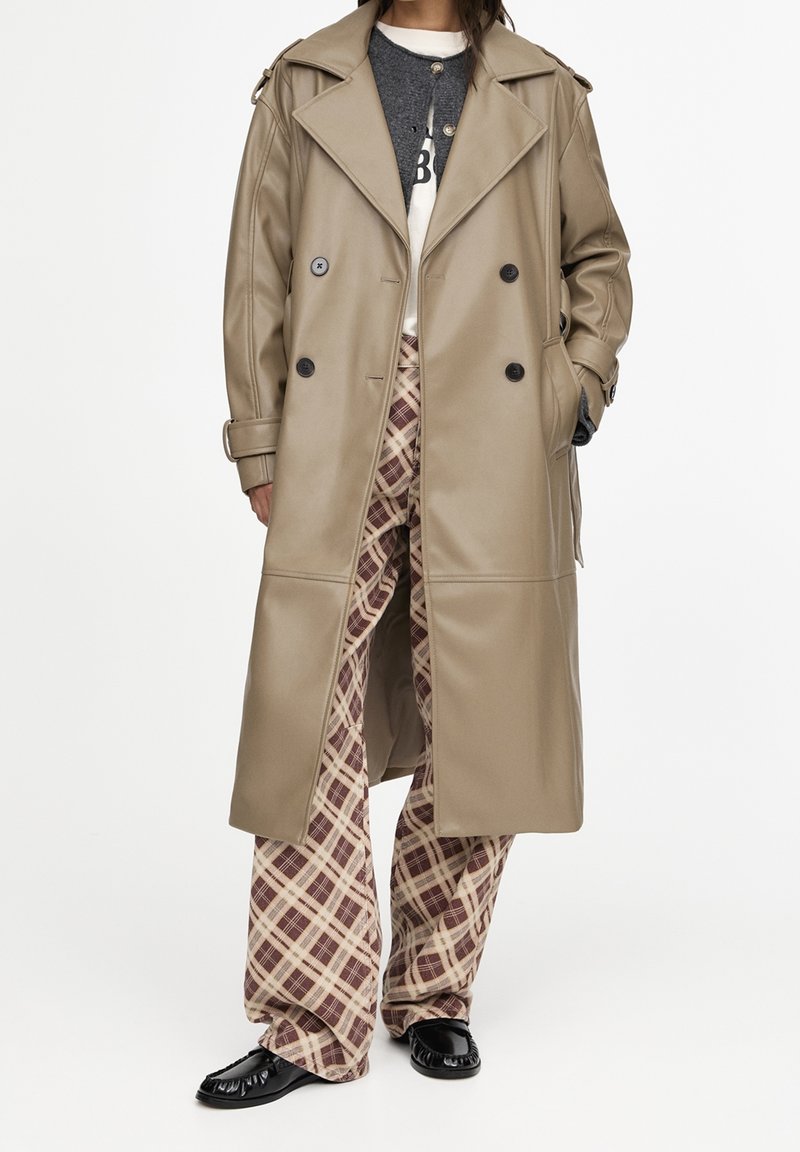 Personne portant un long manteau beige croisé sur un pull sombre et une chemise blanche, associé à un pantalon large beige et marron à carreaux et des chaussures noires.
