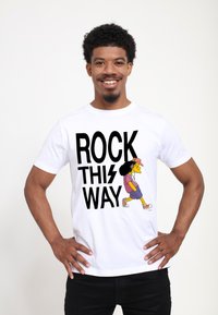 Muž s krátkými kudrnatými vlasy a bradkou, oblečený v bílém tričku s nápisem „ROCK THIS WAY“ a karikaturní postavičkou, která kráčí s sluchátky na uších.