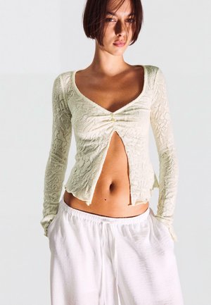 Rövid barna hajú nő, aki krémszínű, részben begombolt hosszú ujjú csipkés felsőt és laza fehér nadrágot visel, kezét a zsebében tartva áll.