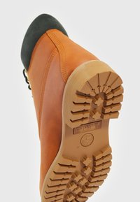 Pomarańczowy skórzany but z czarnym gumowym noskiem, z charakterystyczną chropowatą, brązową gumową podeszwą z wyraźnym wzorem bieżnika.