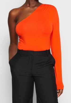 Kobieta w jasnopomarańczowej bluzce na jedno ramię z długim rękawem, w połączeniu z czarnymi spodniami z wysokim stanem, z rękami w kieszeniach.