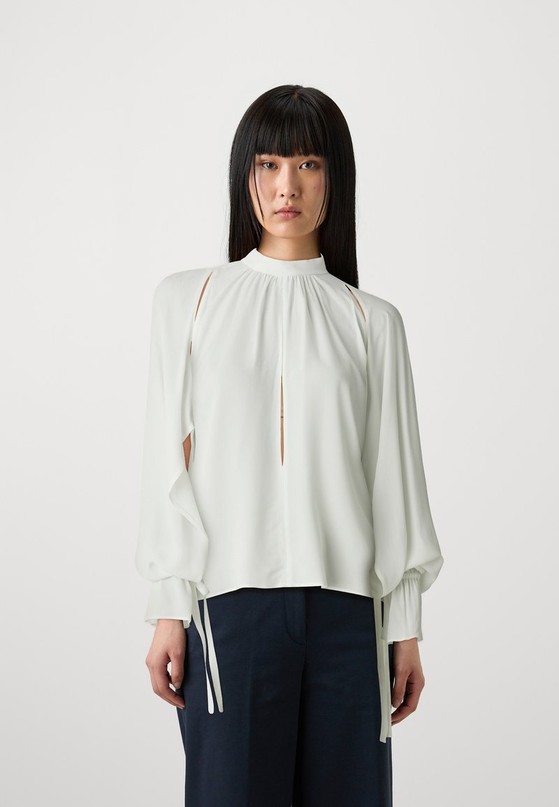 Blusa bianca con un collo alto, maniche lunghe e ritagli laterali. Presenta maniche arricciate con lacci ai polsi e un dettaglio a chiave sul davanti.