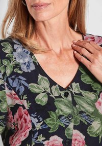 Blouse à motifs floraux en tissu sombre. Caractéristiques : encolure en V, fermeture à boutons, et un mélange de motifs floraux roses, bleus et verts.