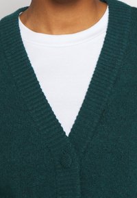 Turkusowy sweter z dzianiny z dekoltem w serek, prążkowanym kołnierzykiem i pojedynczym zapięciem na guzik, założony na biały prążkowany t-shirt.