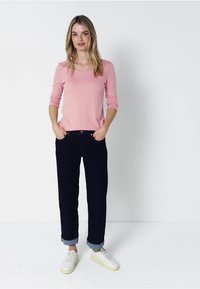 Ljust rosa långärmad tröja, avslappnad passform, ihop med mörkblå jeans med uppvikta ben och vita sneakers. Enkelt design utan synliga mönster eller detaljer.