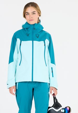 Teal och ljusblå skidjacka med hög krage, framdragkedjor och justerbar huva. Matchas med byxor i samma färg och håller skidglasögon.