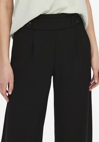 Un pantalon noir sur mesure avec une coupe évasée, présentant un devant plat, deux boutons à la taille et une texture de tissu lisse.