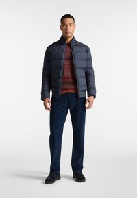 Marineblauwe pufferjack met hoge kraag, gestructureerde stof en doorstikte ontwerpen, gedragen over een geprinte sweater en gecombineerd met donkere corduroy broek.