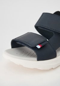 Tamsiai mėlynos sandalai su minkštu guminiu padu, reguliuojamomis velcro juostelėmis ir mažomis raštuotomis detalėmis, sudarytomis iš raudonų, baltų ir mėlynų siūlų.