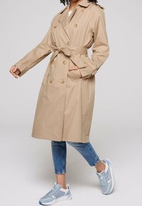 Cappotto trench beige con bottoni doppi, cintura e tasche laterali, indossato sopra jeans blu e sneakers chiare.