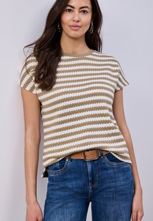 Frau mit langen dunklen Haaren, die goldene Creolen, ein beige-weiß gestreiftes Kurzarmshirt, blaue Jeans und einen braunen Gürtel trägt.