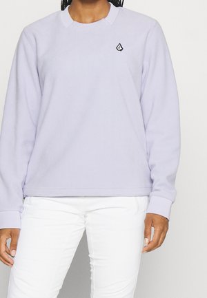 Personne portant une chemise à manches longues de couleur violet clair avec un petit logo noir et blanc sur le côté gauche de la poitrine et un pantalon blanc.