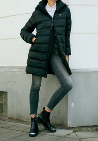 Henkilö, jolla on päällään musta toppatakki, harmaat kapeat farkut ja mustat nilkkurit, seisoo ulkona valkoista ja harmaata seinää vasten.