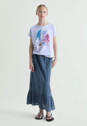 Joven mujer de pie con una camiseta gráfica de color púrpura claro, falda larga azul oscuro con ojales y dobladillo con volantes, y sandalias negras de tiras.