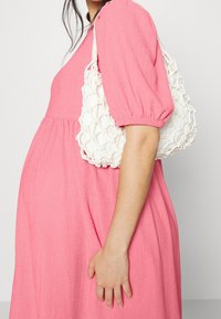 Donna in un vestito strutturato rosa porta una borsa a rete bianca con un sacchetto di stoffa all'interno, mostrata dal collo a metà coscia.