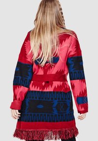 Cárdigan de patrón rojo y negro con acentos azules, que presenta una cintura con cinturón, mangas largas y terminación con flecos, en un tejido de punto texturizado.