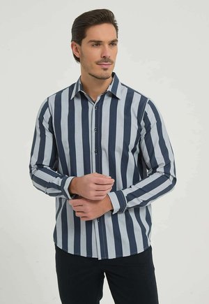 Homme ajustant le poignet d'une chemise à manches longues à rayures verticales bleu marine et gris clair, debout devant un fond clair uni.