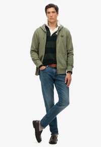 Sudadera verde oliva con bolsillos frontales, sobre una camiseta polo a rayas oscuras. Jeans azules y zapatos marrón oscuros completan el conjunto.