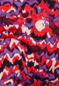 Multicolored fabric featuring a wavy abstract pattern in red, purple, black, and cream. Soft texture with a slightly sheer appearance.