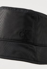 Czarny kapelusz typu bucket wykonany z gładkiego materiału, z usztywnionym rondem, poziomym przeszyciem i haftowanym logo "cK" z boku.