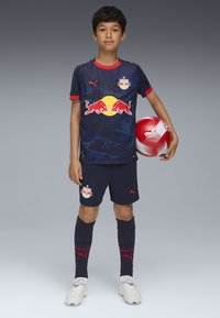 Marintröja med röda detaljer, med en logotyp på tröjan och shorts. Spelaren håller en fotboll med röd och vit mönstring.